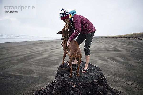 Verspielte Frau steht mit Hund auf Baumstamm am Strand gegen den Himmel