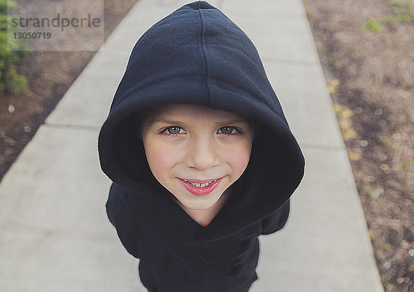 Hochwinkelporträt eines glücklichen Jungen mit schwarzer Kapuzenjacke,  der auf einem Fußweg steht