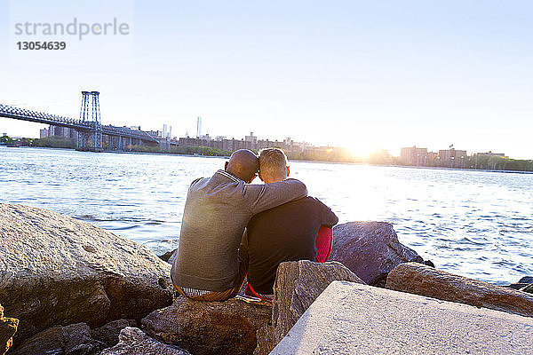 Rückansicht eines Mannes,  der bei Sonnenuntergang mit seinem Freund auf Felsen am East River sitzt