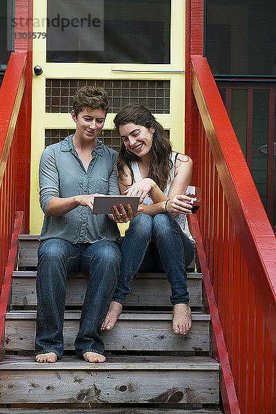 Lesbisches Paar benutzt digitales Tablett,  während es auf einer Treppe vor dem Haus sitzt
