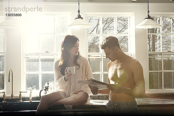 Mann liest Zeitung,  während seine Freundin mit Kaffeetasse auf dem Küchentisch sitzt