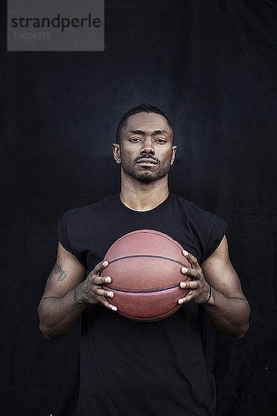Porträt eines Sportlers,  der Basketball in der Hand hält,  während er gegen eine schwarze Wand steht
