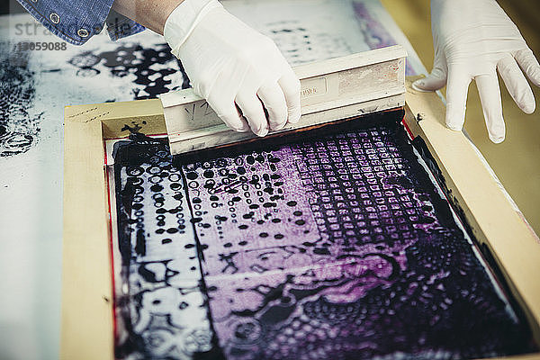 Ausgeschnittene Hände einer Handwerkerin,  die im Siebdruckverfahren das Design in der Werkstatt auf Stoff druckt