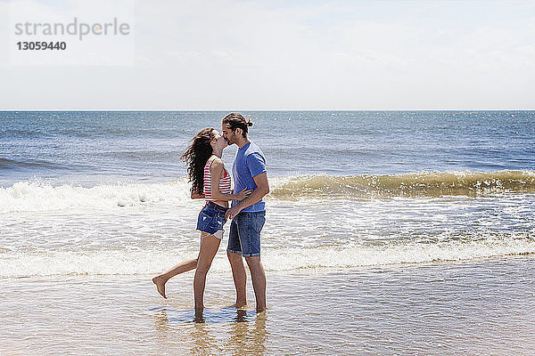 Zärtliches Paar küsst sich,  während es am Strand am Ufer steht