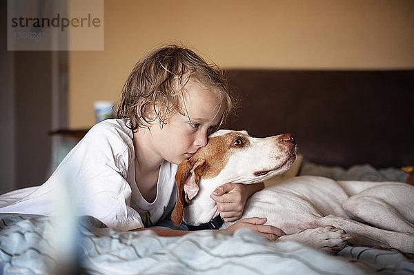 Junge umarmt Hund,  während er zu Hause auf dem Bett liegt