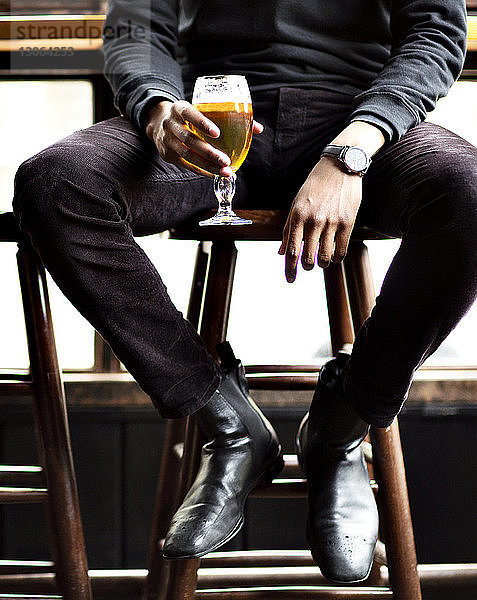 Niedriger Teil eines Mannes,  der Weinglas hält,  während er auf einem Hocker in einer Bar sitzt