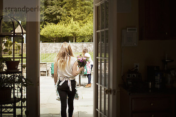 Rückansicht einer Frau,  die eine Blumenvase trägt,  während sie auf dem Rasen geht,  durch die Tür gesehen