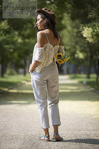 Nachdenkliche Frau hält Sonnenblumen und steht auf einem Fußweg im Park