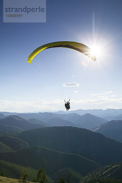 Silhouette einer Person beim Gleitschirmfliegen über Berge vor hellem Himmel