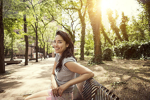 Lächelnde Frau sitzt an sonnigen Tagen im Park auf Bank an Bäumen
