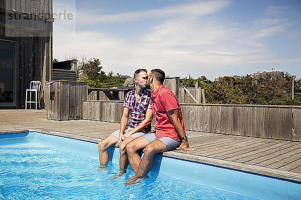 Homosexuelles Paar küsst sich,  während es am Poolrand gegen den Himmel sitzt