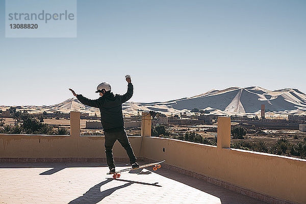 Rückansicht eines Mannes mit Skateboard auf der Gebäudeterrasse vor klarem Himmel