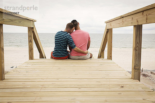 Rückansicht eines homosexuellen Paares,  das auf einer Strandpromenade gegen das Meer sitzt