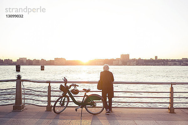 Rückansicht einer älteren Frau,  die bei Sonnenuntergang am Fahrrad gegen das Meer steht