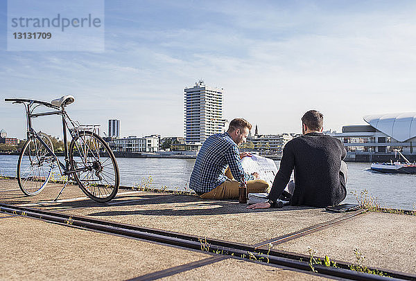 Freunde betrachten Dokument,  während sie mit dem Fahrrad auf der Promenade gegen den klaren Himmel sitzen
