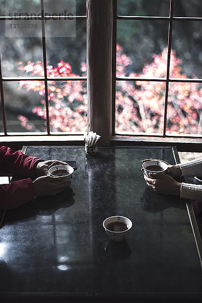 Ausgeschnittenes Bild eines Paares,  das Kräuterteetassen bei Tisch durch ein Fenster hält