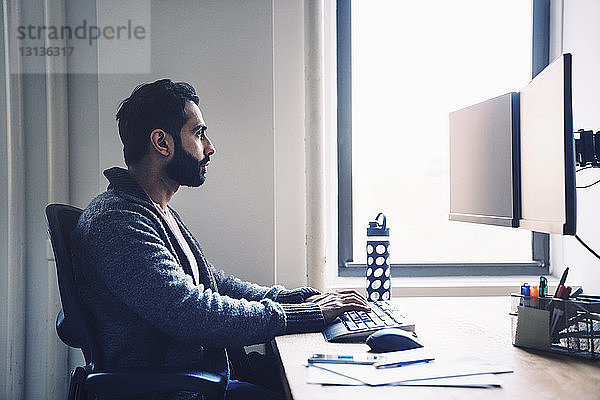Seitenansicht eines Geschäftsmannes,  der einen Desktop-Computer benutzt,  während er im Büro am Schreibtisch sitzt
