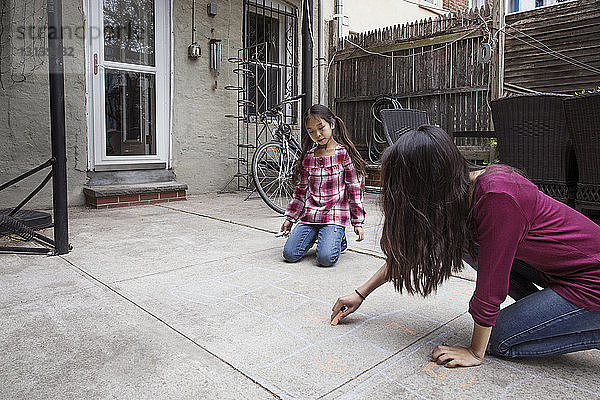Mädchen sieht Schwester an,  die vor dem Haus ein Himmelfahrtskissen zeichnet