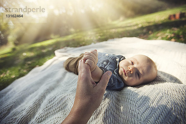 Mutter hält Hand eines kleinen Jungen,  der im Sonnenlicht auf einer Decke liegt und in die Kamera schaut