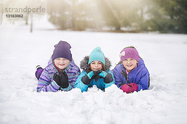 Freunde mit Strickmützen liegen nebeneinander im Schnee auf der Vorderseite und schauen lächelnd in die Kamera