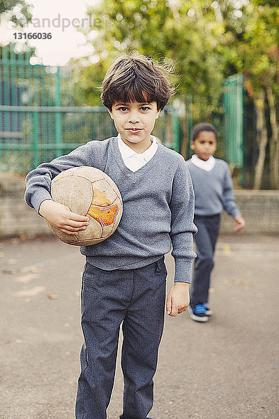 Porträt eines Grundschülers,  der einen Fussball auf dem Spielplatz hält