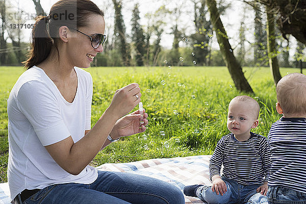 Junge Mutter bläst Blasen für kleine Zwillingsjungen auf Picknickdecke