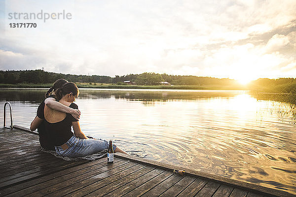 Freundinnen umarmen sich,  während sie bei Sonnenuntergang auf dem Steg über dem See gegen den Himmel sitzen