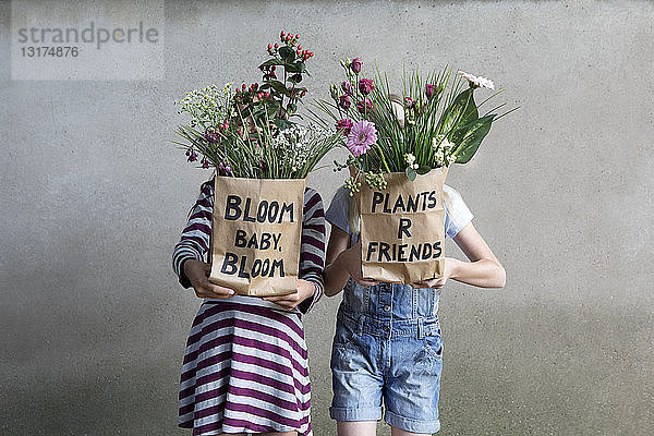 Zwei nebeneinander stehende Mädchen verstecken sich hinter Papiertüten mit Blumen