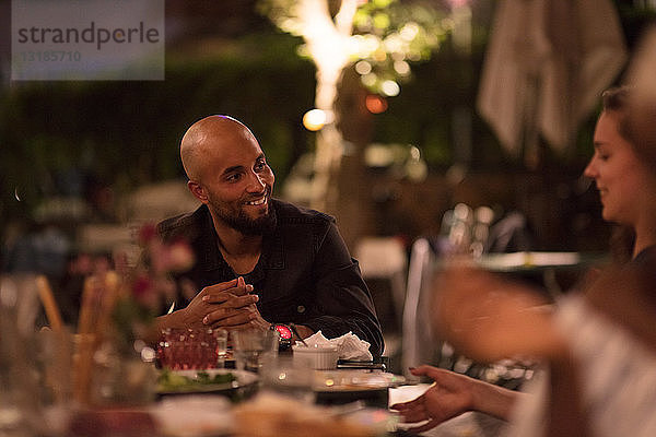 Lächelnder junger Mann mit rasiertem Kopf schaut eine Freundin an,  während er während der Dinnerparty am Tisch sitzt