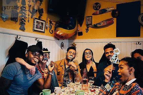 Porträt fröhlicher junger multiethnischer Freunde,  die Requisiten halten,  während sie während der Dinnerparty im Restaurant sitzen