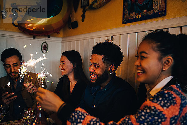 Glückliche multiethnische männliche und weibliche Freunde halten während der Dinnerparty brennende Wunderkerzen im Restaurant
