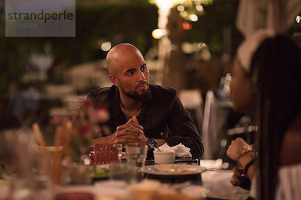 Junger Mann mit kahlrasiertem Kopf schaut eine Freundin an,  während er während der Dinnerparty am Tisch sitzt