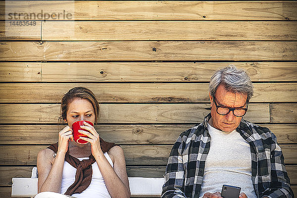 Frau trinkt Kaffee und Mann benutzt Smartphone vor einer Blockhütte