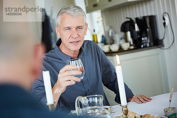 Reifer Mann schaut weg,  während er ein Trinkglas auf dem Esstisch hält,  mit einem Freund im Vordergrund