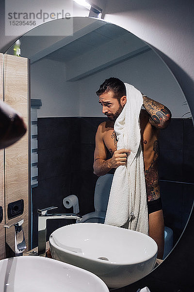 Mittelgroßer erwachsener Mann mit Tätowierungen,  die Haare am Badezimmerspiegel trocknen,  Spiegelbild