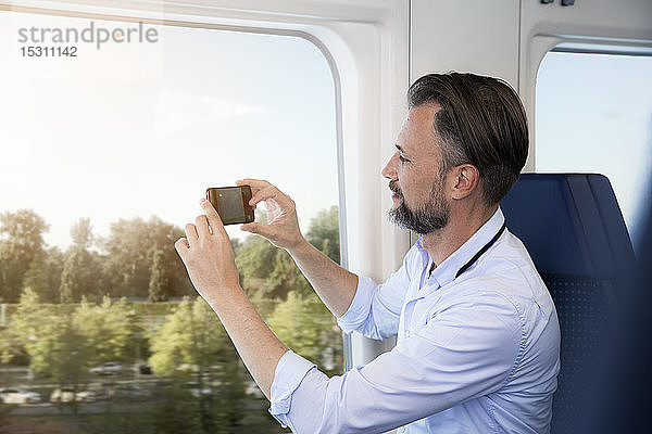 Älterer Mann sitzt in einem Zug und fotografiert mit seinem Smartphone