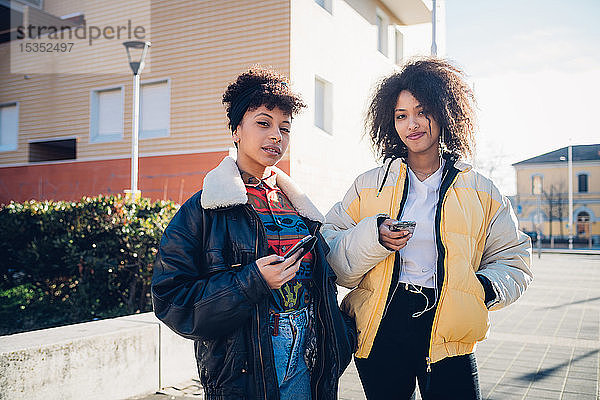 Zwei coole junge Freundinnen mit Smartphones auf dem städtischen Bürgersteig,  Porträt