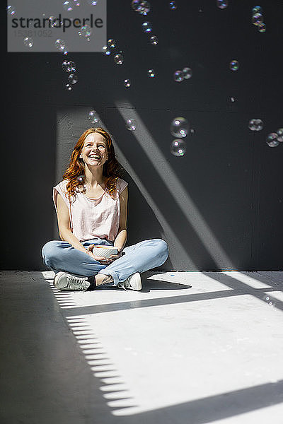 Porträt einer glücklichen Frau,  die mit einer Kaffeeschüssel auf dem Boden sitzt und Seifenblasen betrachtet