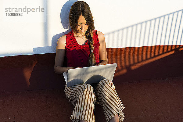 Teenagerin sitzt mit Laptop im Freien im Sonnenlicht