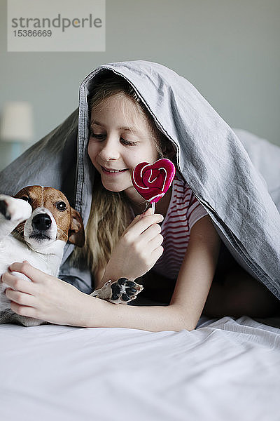 Lächelndes Mädchen liegt auf dem Bett mit einem Lolli,  der ihren Hund kitzelt