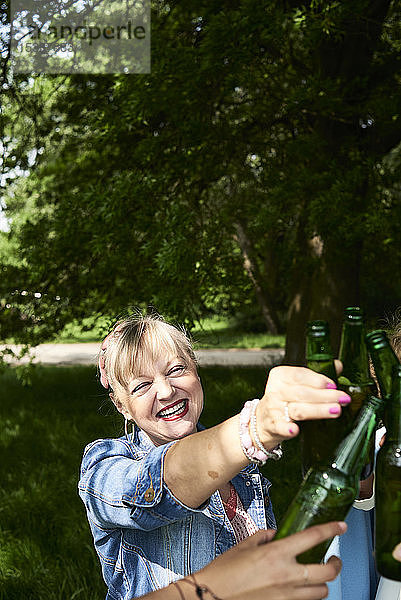 Porträt einer glücklichen Frau,  die mit Freunden im Park an einer Bierflasche klimpert