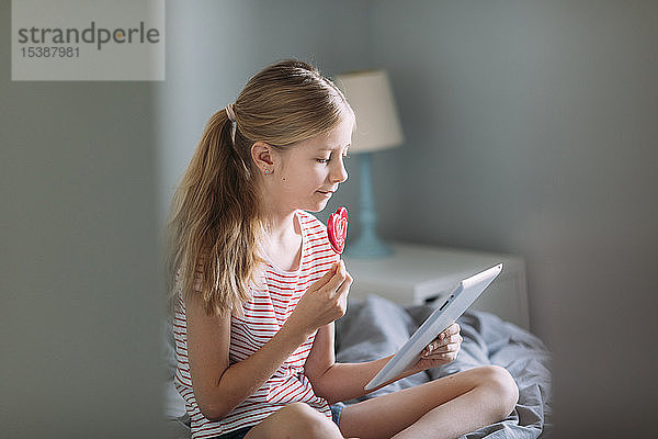 Blondes Mädchen sitzt mit Lolli auf dem Bett und benutzt ein digitales Tablett