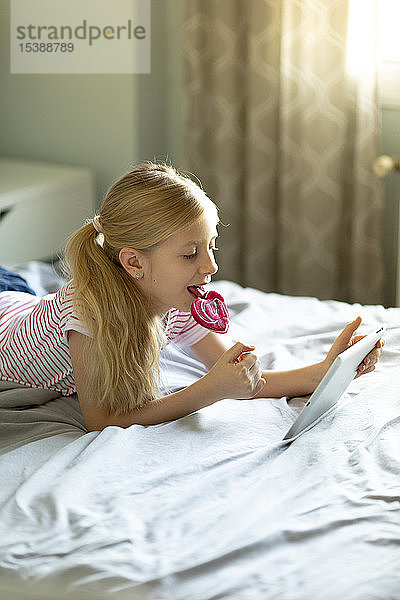 Blondes Mädchen liegt im Bett und leckt Lolli,  während sie ein digitales Tablett benutzt
