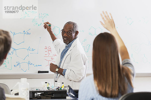 Eine Studentin hebt ihre Hand in einer Ingenieursklasse,  während der Professor an der Tafel über chemische Bindungen schreibt
