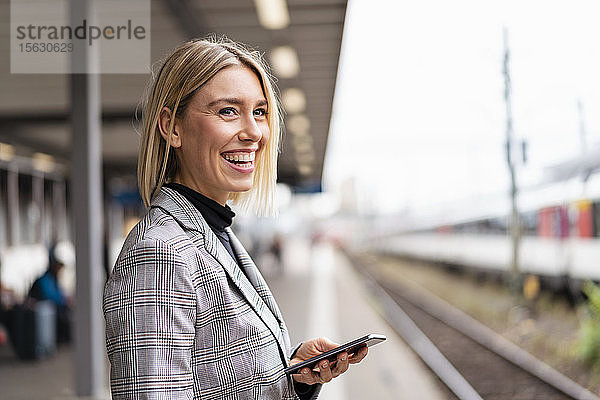 Glückliche junge Geschäftsfrau mit Mobiltelefon am Bahnhof