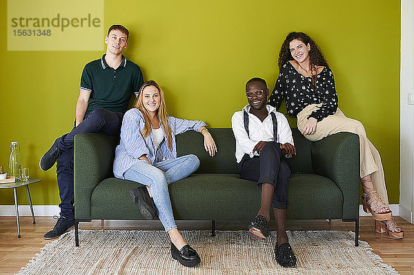 Porträt von lächelnden Kollegen,  die in einer grün ummauerten Bürolounge auf einem Sofa sitzen