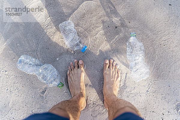 Männerfüße am Strand,  umgeben von leeren Plastikflaschen,  Draufsicht