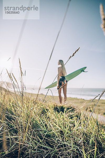 Junge blonde Surferin am Strand,  Fokus auf Gras im Vordergrund