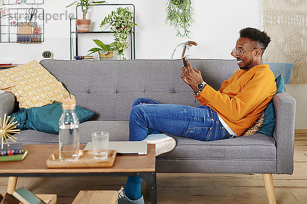 Junger lächelnder Mann sitzt auf einer Couch und benutzt ein Smartphone
