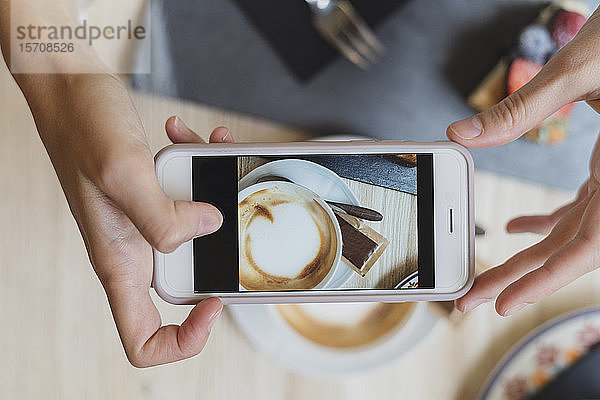 Draufsicht einer Frau in einem Cafe,  die ein Handyfoto von Cappuccino macht
